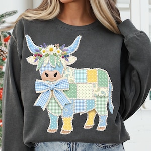 May include: Dark gray sweatshirt featuring a colorful patchwork cow design. The cow has blue horns, a floral crown, and a gingham bow. The patchwork includes blue, green, and yellow squares with paisley and polka dot patterns. A whimsical and unique apparel item.