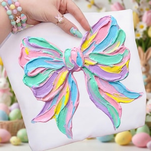 May include: A white pillow with a large, textured bow design. The bow is painted in pastel colors including pink, blue, green, yellow, and purple. The design is a whimsical, artistic representation.