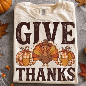 Op de afbeelding: Een crèmekleurig t-shirt met de tekst GIVE THANKS in een verweerd donkerbruin lettertype. Het ontwerp bevat een centrale illustratie van een kalkoen met een bruine hoed, geflankeerd door twee pompoenen met oranje en bruine ruitpatronen.