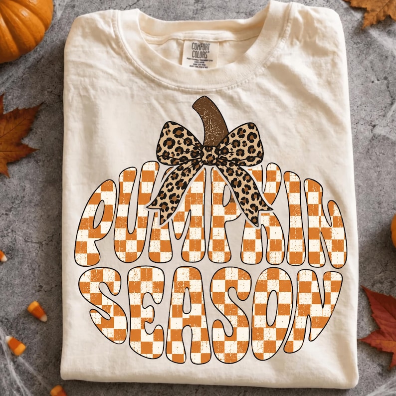 May include: A cream-colored short-sleeve t-shirt featuring a large pumpkin graphic filled with an orange and white checkered pattern. The pumpkin is topped with a brown stem and a leopard print bow, with the text PUMPKIN SEASON in bold letters.