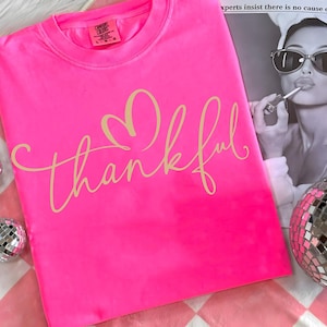 May include: Bright pink Comfort Colors crew neck sweatshirt with the word "thankful" in a cursive font and a heart above the word. The sweatshirt is on a pink and white checkered surface.