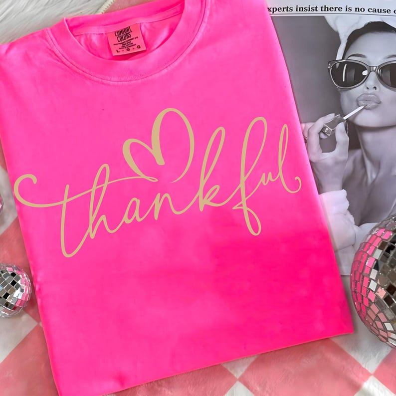 May include: Bright pink Comfort Colors crew neck sweatshirt with the word "thankful" in a cursive font and a heart above the word. The sweatshirt is on a pink and white checkered surface.