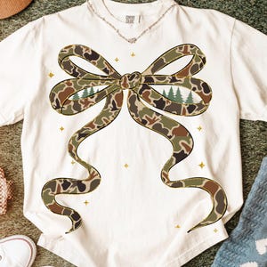 May include: White t-shirt featuring a large bow design in camouflage pattern with gold trim. The bow has a forest scene in the center. The shirt has the words "Comfort Colors" printed on the collar.