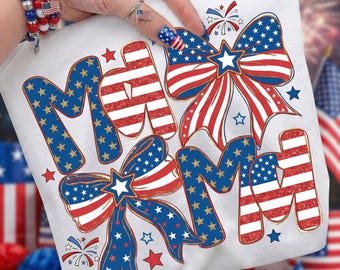Op de afbeelding: Wit T-shirt met een patriottisch ontwerp. Het woord "MOM" wordt weergegeven in rood-witte strepen en blauwe sterren. Twee strikken met rood-witte strepen en blauwe sterren zijn ook aanwezig. Vuurwerk en sterren maken het ontwerp compleet.