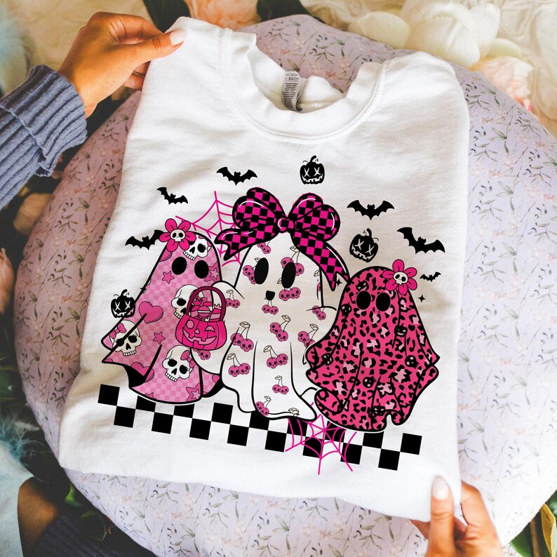 May include: White sweatshirt featuring three ghost characters with pink and black accents. The ghosts have various patterns, including skulls, cherries, and leopard print. The design includes bats, spiderwebs, and a checkered border, perfect for Halloween.