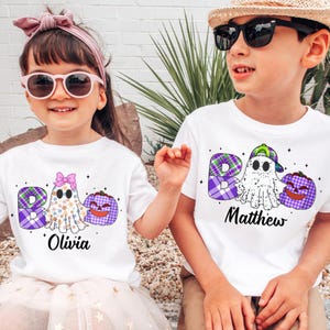 May include: Two children wearing white t-shirts with Halloween designs. The shirts feature the word "BOO" in a plaid pattern, a ghost, and a jack-o'-lantern. One shirt has the name "Olivia", the other has "Matthew".