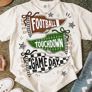May include: A white t-shirt featuring three pennant-style graphics. The top brown pennant reads FOOTBALL, the middle green pennant reads TOUCHDOWN with yard line markings, and the bottom black and white checkered pennant reads GAME DAY.