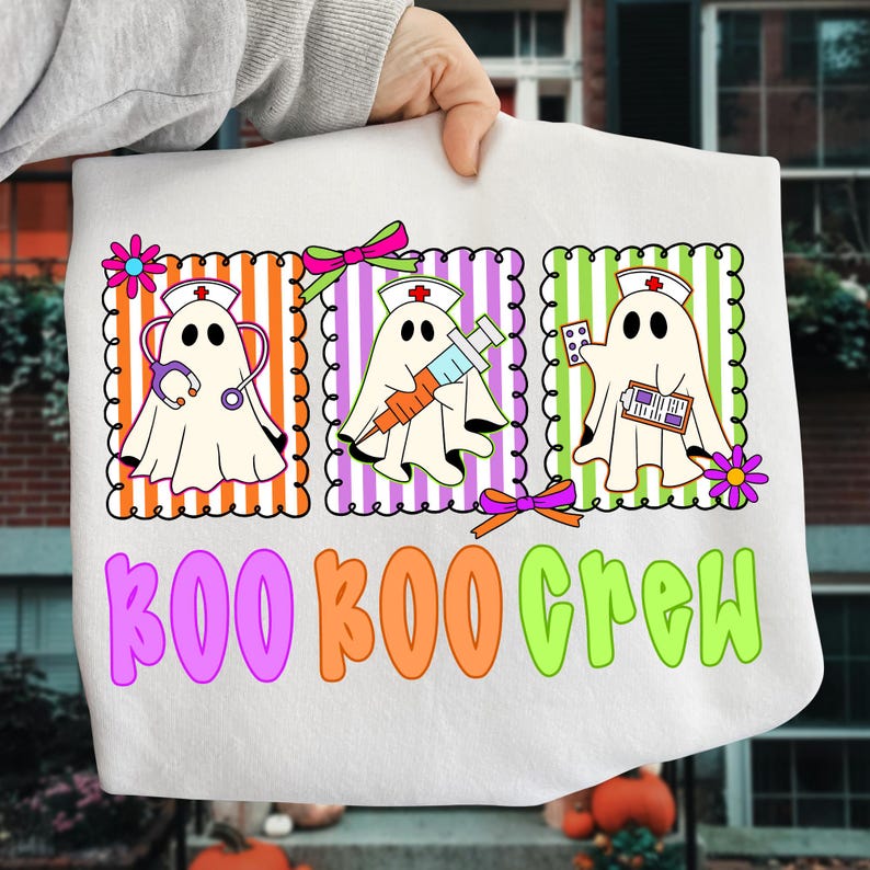 May include: White Halloween-themed graphic tee featuring three ghost characters dressed as nurses, each with medical tools. The text "BOO BOO CREW" is in colorful bubble letters. The design includes orange, purple, green, and pink accents.