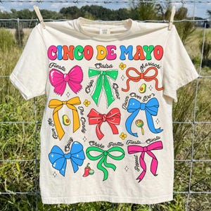 May include: Off-white t-shirt with a colorful Cinco de Mayo design. The graphic includes the text "Cinco de Mayo" in rainbow colors, along with various bow illustrations and food-related words like "Fiesta" and "Salsa".