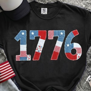 Może przedstawiać: Czarny t-shirt z numerem "1776" w patchworkowym wzorze. Numery wykonane są z czerwonych, białych i niebieskich tkanin, w tym dżinsu, kraty i wzorów kwiatowych. Każda liczba ozdobiona jest białą gwiazdą.
