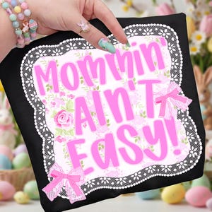 Puede incluir: Camiseta negra con la frase rosa "Mommin' Ain't Easy!" rodeada de un diseño floral y un borde de encaje blanco. Lazos rosas adornan la camiseta. Un regalo divertido para madres.
