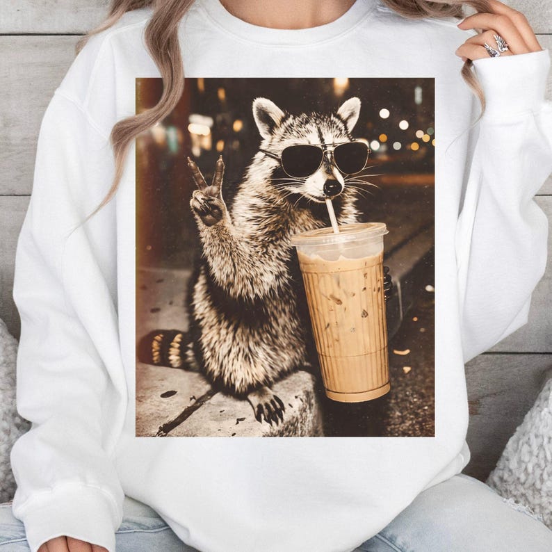 May include: White sweatshirt with a raccoon wearing sunglasses, holding an iced coffee, and giving a peace sign. The image has a sepia tone, with a blurred background of city lights.