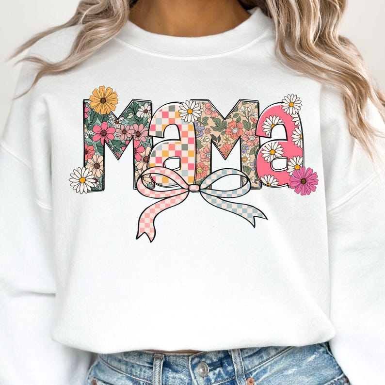 May include: White sweatshirt with the word "MAMA" in a floral design. The letters are filled with various floral patterns, a checkered pattern, and daisies. A bow with pink and blue checkered ribbons is below the word.