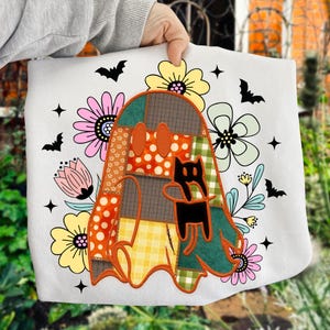 May include: A white sweatshirt featuring a central graphic of a ghost made of various orange, green, and brown patterned fabric patches. The ghost holds a small black cat. The design is surrounded by colorful flowers, black bats, and stars.
