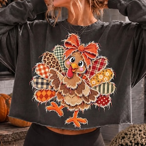 May include: A charcoal gray long-sleeve sweatshirt featuring a central graphic of a turkey with a large orange bow. The turkey's feathers are depicted as a collage of various patterns including leopard print, plaid, gingham, and floral designs.