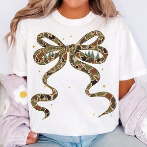 May include: White t-shirt featuring a large bow design in a camouflage pattern with brown, green, and tan hues. The bow has a forest scene in the center. Gold star accents are scattered around the bow.