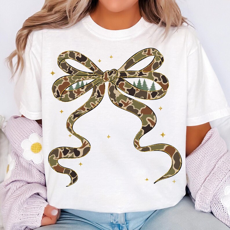 May include: White t-shirt featuring a large bow design in a camouflage pattern with brown, green, and tan hues. The bow has a forest scene in the center. Gold star accents are scattered around the bow.
