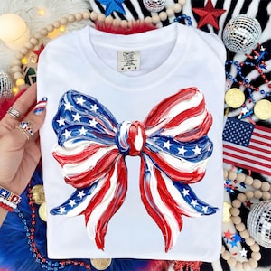 May include: White t-shirt featuring a large, textured bow design in red, white, and blue, with stars. The bow is painted to resemble the American flag. The shirt has a tag that says "Comfort Colors".
