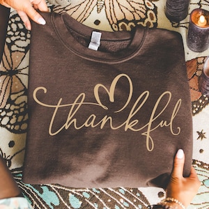 May include: A brown sweatshirt with the word "thankful" in a cursive font and a heart design. The sweatshirt is held by two hands. The background includes a patterned cloth and candles.