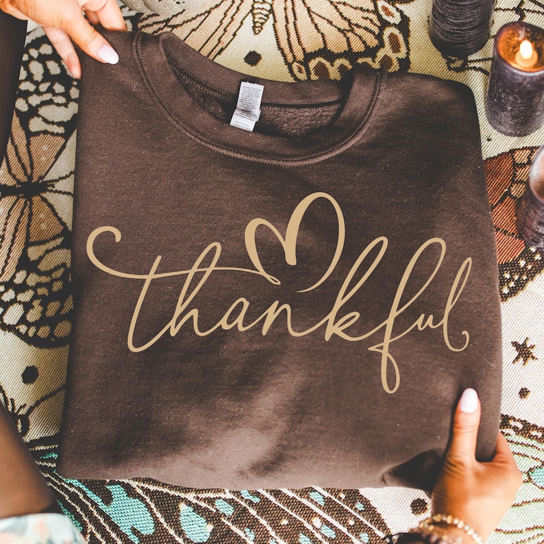 May include: A brown sweatshirt with the word "thankful" in a cursive font and a heart design. The sweatshirt is held by two hands. The background includes a patterned cloth and candles.