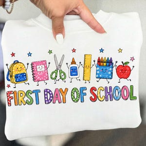 Puede incluir: Una sudadera blanca con un gráfico colorido de útiles escolares animados, incluyendo una mochila, cuaderno, tijeras, pegamento, regla, crayones y una manzana. El texto "FIRST DAY OF SCHOOL" está impreso en letras multicolores y estampadas.