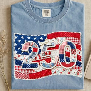 Puede incluir: Camiseta azul claro con un diseño patriótico. El diseño presenta una bandera estadounidense de patchwork con el número "250" en el centro. La bandera está hecha de tela estampada roja, blanca y azul, y tiene un lazo blanco en la parte superior.