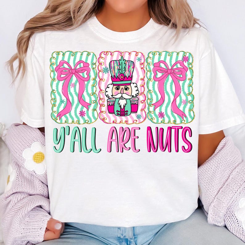 以下が含まれることがあります： カラフルなグラフィックデザインが施された白いTシャツ。デザインには、2つのピンクのリボンとくるみ割り人形の兵士の3つのフレーム要素が含まれています。「Y'ALL ARE NUTS」のテキストは、フレーム要素の下にティールとピンクで印刷されています。