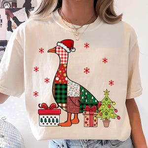 Puede incluir: Una camiseta color crema con el gráfico de un ganso que lleva un gorro de Papá Noel rojo y blanco. El cuerpo del ganso tiene un patrón de retazos de cuadros y flores. Está rodeado de copos de nieve rojos, dos regalos y un pequeño árbol.