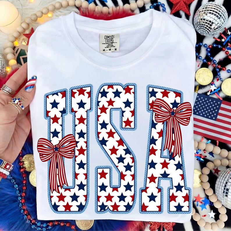 May include: White t-shirt with the word "USA" in large letters. The letters are filled with red and blue stars and outlined in blue. Red and white bows are on the letters "U" and "A". The shirt has a tag that says "Comfort Colors".
