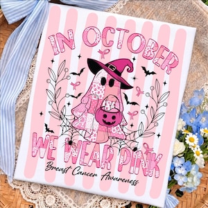 May include: A rectangular sign featuring a pink and white striped background with a ghost wearing a witch hat and holding a pumpkin bucket. The text reads IN OCTOBER WE WEAR PINK Breast Cancer Awareness, surrounded by bats, webs, and ribbons.