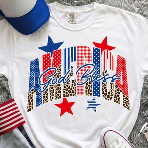 Könnte beinhalten: Weißes T-Shirt mit dem Wort "AMERICA" in rot, weiß und blau gemustert, und den Worten "God Bless" in Schreibschrift. Das Design beinhaltet Sterne und ein Leopardenmuster. Eine blau-weiße Baseballkappe und eine kleine amerikanische Flagge sind sichtbar.