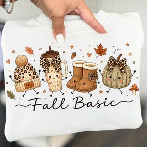 May include: A white sweatshirt featuring a graphic print of a cartoon beanie, a tumbler, boots, and a pumpkin, all with smiling faces. The design includes leopard print accents, autumn leaves, and the text Fall Basic in a black cursive script.