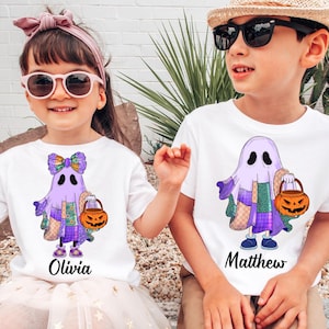 May include: Two children wearing white t-shirts with a cartoon ghost design holding a jack-o'-lantern. The ghost is purple with patchwork details. One shirt says "Olivia" and the other says "Matthew".
