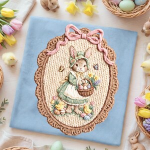 以下が含まれることがあります： イースターをテーマにした刺繍の壁掛け。ボンネットをかぶったウサギが、卵と花が入ったバスケットを持っています。デザインはスカラップの縁とピンクのリボンで縁取られ、水色の背景に配置されています。画像には、ひよこ、蝶、イースターエッグが含まれています。