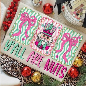 May include: A beige t-shirt with the text "Y'all Are Nuts" in teal and pink. The shirt features three decorative panels with pink bows, snowflakes, and a nutcracker design. Christmas-themed apparel.