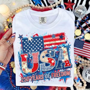 May include: White t-shirt with a patriotic design featuring the American flag and the word "USA" in red, white, and blue patchwork. The text "250 YEARS OF FREEDOM" is below the graphic. The shirt is a Comfort Colors brand.