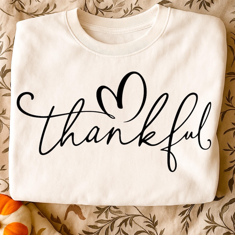 May include: Cream-colored sweatshirt with the word "thankful" in black script, featuring a heart-shaped design above the word. The sweatshirt has a crew neck and long sleeves, perfect for fall and winter.