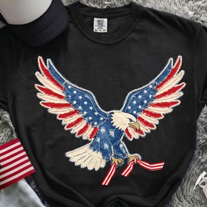 May include: Black t-shirt featuring a patriotic design of an eagle with outstretched wings. The eagle's wings are adorned with a denim-like pattern, incorporating red and white stripes and blue with white stars. The shirt also has the text "COMFORT COLORS" and "EL-75-80".