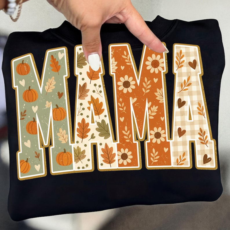 May include: A black sweatshirt featuring the word MAMA in large block letters. Each letter has a unique fall-themed pattern including orange pumpkins on sage green, autumn leaves on cream, sunflowers on orange, and a beige buffalo check.