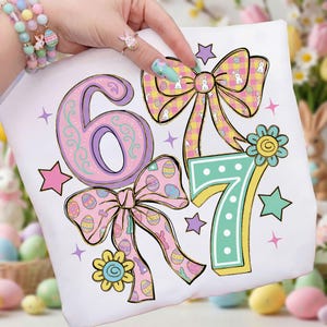 Pode incluir: Camiseta branca com os números 6 e 7 em cores pastel, decorada com laços, estrelas e flores. Os laços apresentam motivos de ovos de Páscoa e coelhinhos. Perfeita para a Páscoa ou um aniversário.