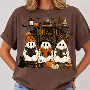 May include: A brown t-shirt featuring a graphic of three white ghosts reading books in front of a bookshelf. The ghosts wear a beanie, a witch hat, and a plaid bow. The design includes pumpkins, a black cat, candles, and the text Curses & Hexes.