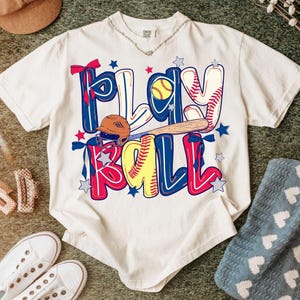 Op de afbeelding: Crèmekleurig T-shirt met de tekst "PLAY BALL" in een kleurrijk honkbalthema-ontwerp. Het ontwerp bevat een honkbal, een knuppel, een helm en sterren. Andere items, waaronder schoenen en een hoed, zijn zichtbaar.