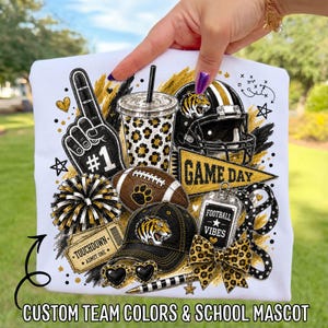 May include: A white t-shirt featuring a graphic print of football-themed items including a number one foam finger, a leopard print tumbler, a tiger mascot helmet, a football, a pennant reading GAME DAY, a ticket, a cap, and a bow.