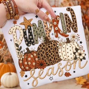 May include: A white t-shirt featuring the text PUMPKIN season in a decorative font. The letters and three pumpkins are filled with various patterns including leopard print, polka dots, and floral designs, accented with small stars and flowers.