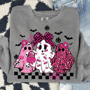 May include: Gray sweatshirt featuring three ghost characters with pink and black accents. The ghosts have various patterns, including cherries, leopard print, and checkered bows. Halloween-themed design with bats and pumpkins.