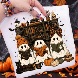 May include: A white t-shirt featuring a graphic of three ghosts reading books in front of a bookshelf. The ghosts wear an orange beanie, a black witch hat, and a plaid bow. The design includes pumpkins, a black cat, a skull, and the text Spells, Potions, Curses, and Hexes.