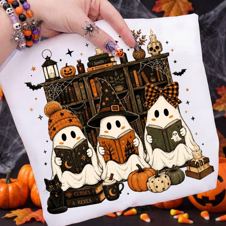 May include: A white t-shirt featuring a graphic of three ghosts reading books in front of a bookshelf. The ghosts wear an orange beanie, a black witch hat, and a plaid bow. The design includes pumpkins, a black cat, a skull, and the text Spells, Potions, Curses, and Hexes.