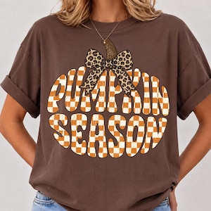 May include: A dark brown short-sleeved t-shirt featuring a large graphic of a pumpkin shape filled with an orange and white checkered pattern. The text PUMPKIN SEASON is written in white across the pumpkin, topped with a leopard print bow.