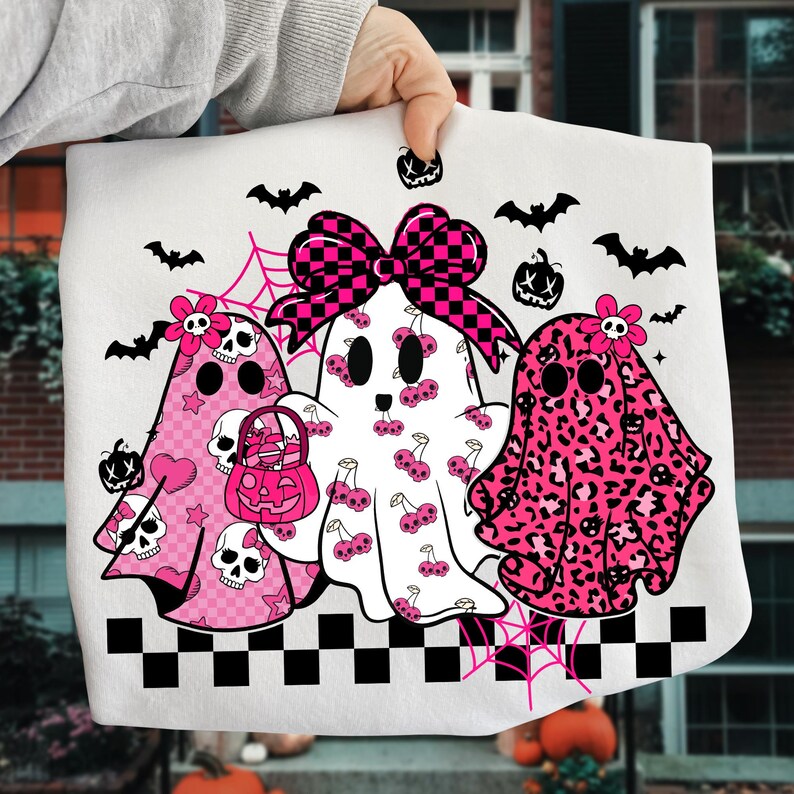 May include: White Halloween-themed tote bag featuring three ghost characters with pink accents. The ghosts have various patterns, including cherries, leopard print, and hearts. Bats, pumpkins, and a checkered bow complete the design.