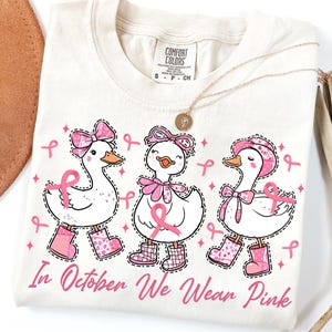 Puede incluir: Camiseta color crema con tres patos de dibujos animados con lazos, cintas y botas rosas. El texto "In October We Wear Pink" está impreso en rosa. Un collar dorado con un colgante está sobre la camiseta.